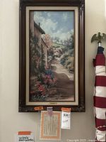 Framed print showing a pathway scene with English cottages and flowers, with a certificate of authenticity hanging below.