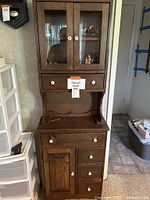 Front view of tall shallow wood cabinet with glass doors on the top, single drawer under the glass doors, open area under drawer, lower cabinet door on left and three drawers on right. Shows wear and has white round knob pulls.