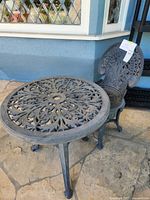 Photo of a small outdoor metal cafe table and metal chair set with intricate floral and scroll designs. Set is on stone patio in front of blue and white building.