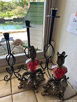 Photo shows 2 ornate metal candlesticks with red faceted glass and 3 dark green metal spiral design candlesticks on a tiled window sill.