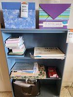 Light blue three-shelf unit containing a blue floral box, box of 25 all occasion greeting cards, notebooks, papers, and a black bag with stationery.