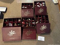 Five burgundy compartmentalized Danbury Mint storage boxes filled with assorted Christmas ornaments on carpeted floor.