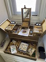 Opened gold-tone jewelry box showing mirror, top trays and assortment of earrings, necklaces, brooches