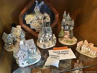 Six English cottage figurines grouped together on a glass shelf with a sign stating 'David Winter Cottages'. Various styles including timber framing and turreted tower design.
