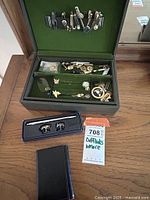 Green velvet-lined jewelry box opened showing multiple cufflinks and tie bars inside and attached to lid, with black wallet in front and boxed pen and cufflinks on side.