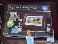 Photo of the Sylvania 10" digital photo frame box showing front with image of frame, memory size, clock/calendar display, and remote control illustration.