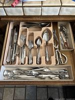 Top view of drawer containing beige plastic flatware sorting tray with assorted knives, forks, spoons, serving utensils, metal tongs, and scissors. Below is a white tray holding additional spoons.