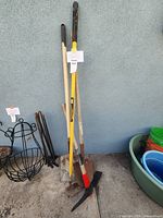 Gardening tools including a pickaxe with yellow handle, a shovel with wooden handle and red blade, spade with wooden handle, black metal plant stands, and colorful plastic containers