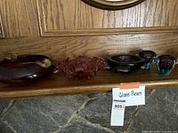 Wide front view of all glass pieces showing dark red, pinkish, blue, and purple colored heavy glass items lined on wooden surface