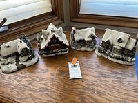 Four Thomas Kinkaid holiday village houses placed on a wooden floor near a window, displaying varying sizes and designs with snow-covered roofs and holiday details.