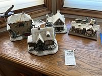 Four holiday village houses displayed on wooden surface, showing snow and Christmas decorations on roofs and bushes. Houses appear dusty and slightly dirty from garage storage.