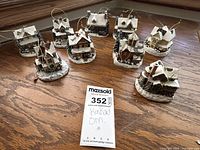 Nine miniature Thomas Kinkaid holiday village ornaments shown on wood surface. Each designed as a snowy small house, hanging loops visible for ornament use.
