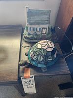 Side photo of a small ceramic house and a stained glass turtle lamp, both placed on a wooden table with cords visible for light-up functionality.