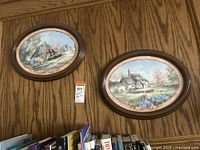Two oval framed artist prints hanging on a wood paneled wall, showing cottage and village scenes with flower gardens.