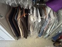Overview of men's jackets and pants hung in a closet space, showing various fabrics and colors, some covered in plastic dry cleaning bags.