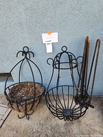 Two black metal hanging planters placed on concrete floor against grey wall. Left planter has coconut coir liner, right planter has decorative cage shape with hook.