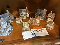 Wide shot showing full set of 10 assorted English style cottage figurines displayed together on a wooden surface including a note card identifying them as David Winter cottages.
