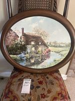 Framed oval print titled "Murrie Cottage" depicting a countryside cottage scene near a waterway displayed on a chair.