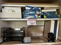 Office supplies including boxes of printer paper, envelopes, blue File-Pro hanging file folders, black plastic paper trays, white binders, and pencil sharpeners on a shelf.