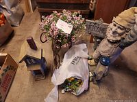 Overall view of gnome, birdhouse, metal flower stand with pink flowers, and dried flower bunches in a plastic bag