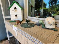 White and green wooden birdhouse alongside three turtle figurines and metal sundial on tiled outdoor surface near window.