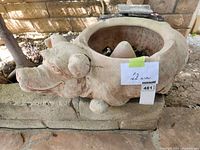 Image of a pig-shaped garden hose holder with a hollow center for hose storage. Pig is off-white color, placed against brick wall outdoors.