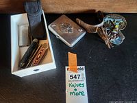 Overview of all lot items on table: pocket knives, lighter, black belt holster pouch, screw driver, two decorative metal belt buckles.