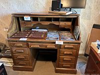 Roll-top desk open with view of inner small drawers, pigeonholes, and larger bottom drawers, showing desk surface with items on it.
