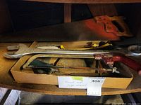 Photo shows a collection of four hand tools in a cardboard box: a wood-handled handsaw, a yellow and black hacksaw, a red handled bow saw, and a large metal open-end wrench.