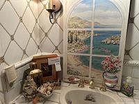 Shows assortment of seashell items on a sink counter including seashell dish, framed seashell, brass lantern, and wall hanging of a scenic window to the sea.