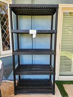 Front view of a black heavy duty plastic shelving unit with five shelves, measuring approximately 35x73x17 inches, placed outside near window and door.