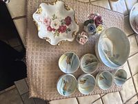 Overview of all china pieces on a towel including large bowl, six small scenic bowls, small tray, and two floral porcelain flower pots