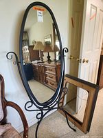 Photo of oval standing mirror with black metal frame and curled ornate stand, showing room reflection.
