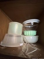Multiple vintage plastic kitchen storage containers and a cake carrier with handle inside a wooden cabinet.