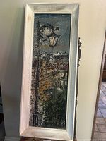 Full view of the vertical painting depicting a street lamp and surrounding buildings framed in a distressed white wooden frame.