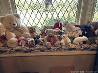 Wide shot of a collection of stuffed animals including various teddy bears, dogs, and other animals arranged on a cushioned window seat.