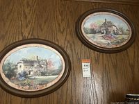 View of both oval framed prints hanging on a wood-paneled wall, showing their size and framing style.