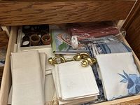 Drawer view showing folded blue and white linens, white napkins with embroidery, brass napkin rings, and another boxed item.