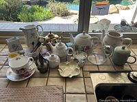 Overview of all teapots, cups, strainers, trays arranged on a kitchen counter with tile surface and window background