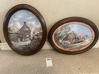 Two oval framed cottage prints by Marty Bell placed on carpeted floor against wall, showing both artworks clearly with wood frames.
