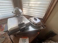 Photo showing four vintage kitchen items: drink dispenser (glass bowl with metal base), covered serving dish, waffle iron, and warming tray, all on a wooden surface.