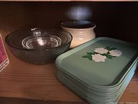 Shelf with 3 clear glass bowls nested inside each other, 1 ceramic bowl behind them, and a stack of light green vintage metal trays with white floral decoration