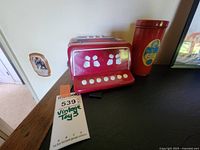Red vintage toy accordion with white buttons and floral decorative accents, placed next to a red cylindrical can with a colorful label indicating snake in a can toy.