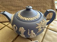 Side view of blue Jasperware Wedgwood teapot with white relief depicting classical figures and foliage.