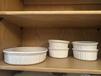 Photo of four smaller white round bowls with ribbed/fluted sides and one clear plastic cover, placed side by side on a wooden shelf.