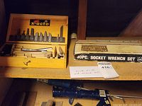 Photo showing wooden X-Acto cutter box with blades mounted inside lid, handles and blades in the box, and socket wrench set box on shelf.