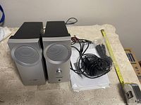 Front view of Bose silver computer speakers with volume control knob on one speaker, power adapter, cables, user manual, and black webcam placed on a table