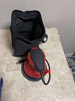 Overall view of the red and black palm style electric car wax buffer on a table with its black carrying bag.