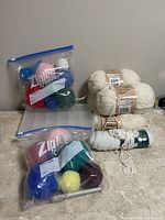 Photo showing two Ziploc bags filled with assorted colorful yarn balls and four skeins of chunky white yarn and one white textured yarn skein on a countertop.