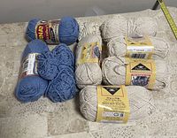 Photo showing a total of 11 yarn skeins, 4 blue (2 new, 2 partial use) and 7 off-white skeins (mostly new) placed on a beige patterned fabric surface.